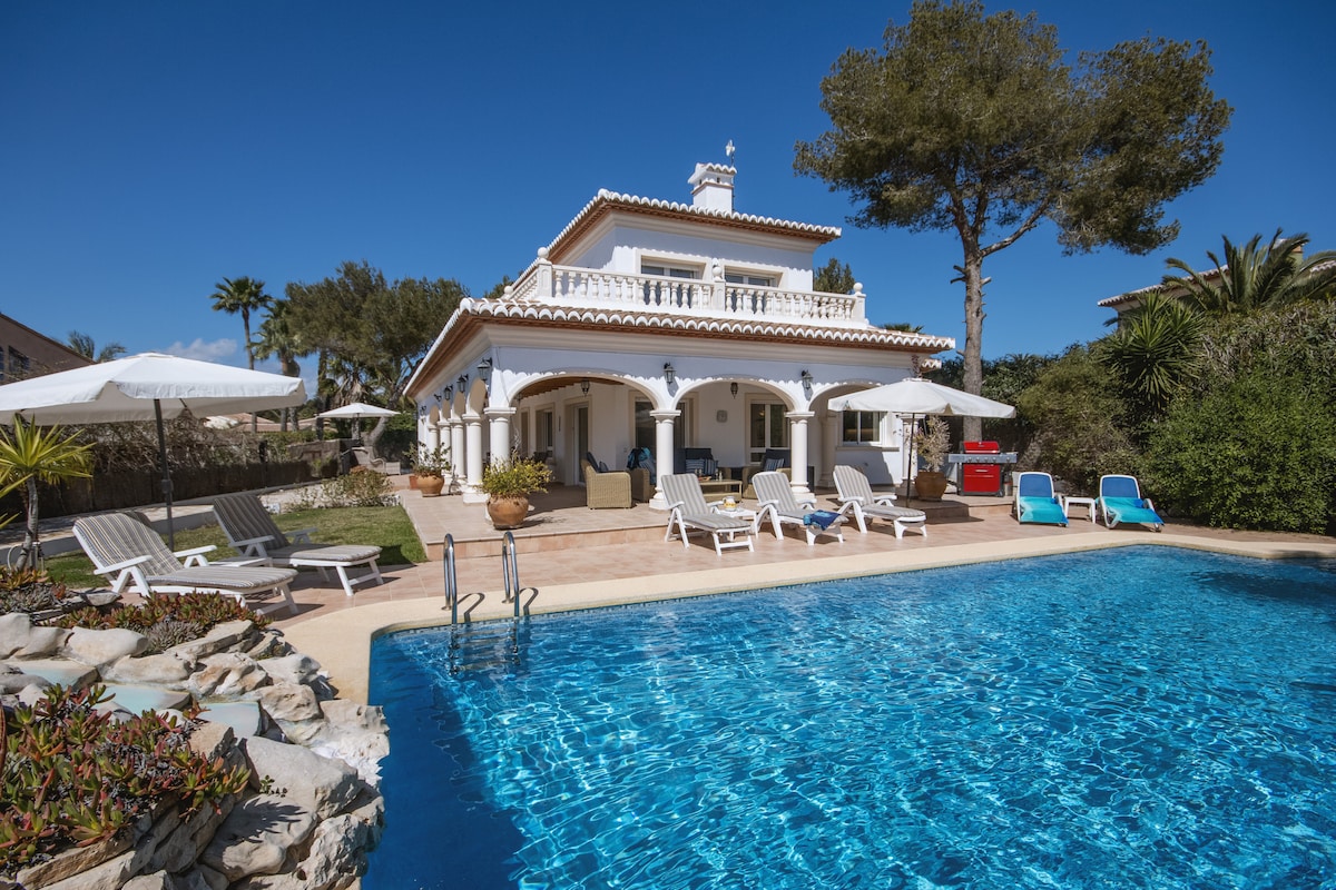 The villa features a spacious outdoor area highlighted by a large swimming pool surrounded by sun loungers and umbrellas. The two-story structure is adorned with white columns and has a terrace overlooking the pool. Lush greenery and palm trees are visible throughout the property.