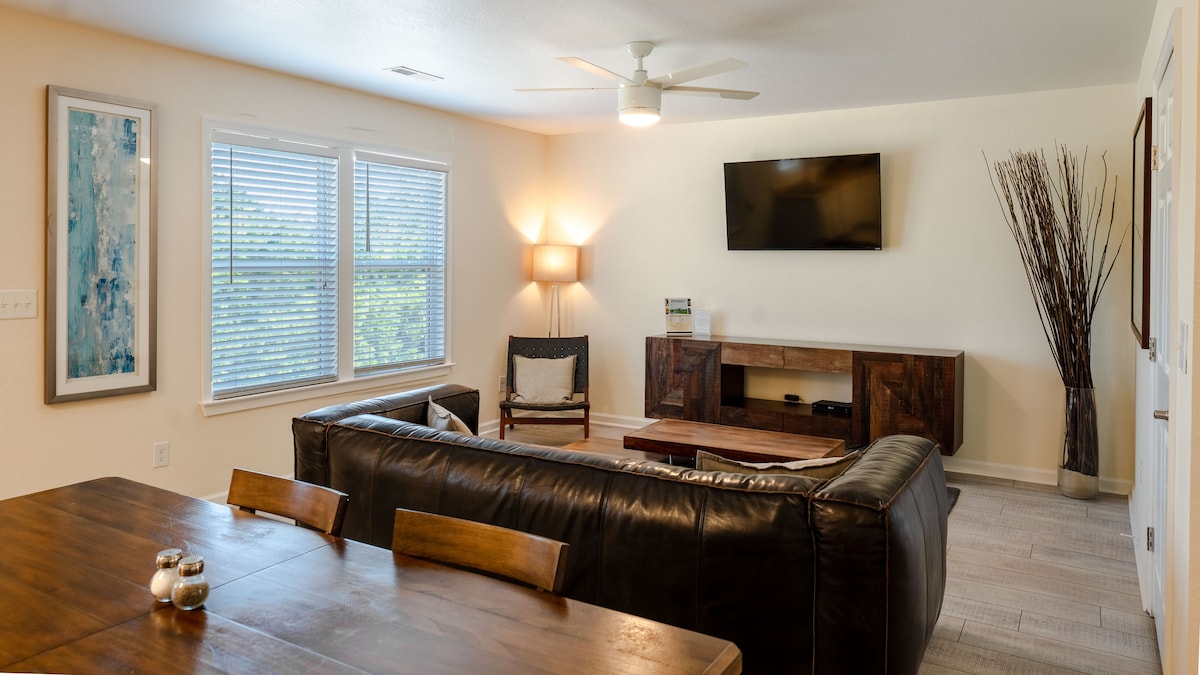 The living area features leather furniture arranged around a wooden coffee table. A smart TV is mounted on the wall opposite a console table. Natural light enters through two large windows with blinds, complemented by a floor lamp and decorative plants.