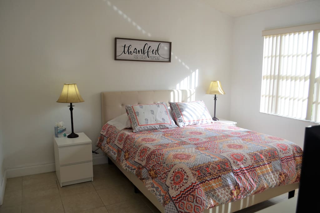 A bright bedroom features a spacious bed adorned with a patterned quilt. Two bedside lamps provide soft lighting on either side. A window with vertical blinds allows natural light to fill the room, and a wall-mounted sign displays a message of gratitude.