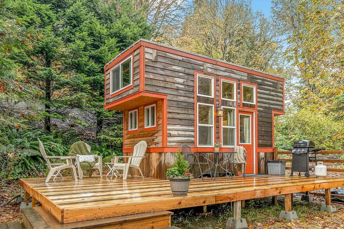 A charming tiny house is nestled among lush green trees, featuring large windows that invite natural light. A wooden deck extends outward, furnished with several chairs and a table, while a grill is positioned nearby for outdoor cooking.
