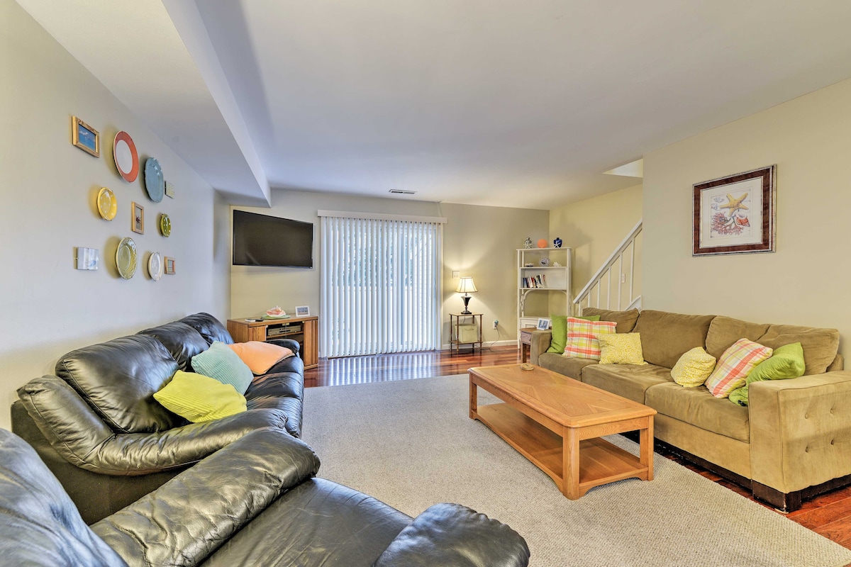 A spacious living area features a combination of black leather sofas and a tan fabric couch, arranged around a wooden coffee table. Light-colored walls are accented by decorative plates and framed artwork. A large sliding door allows natural light to fill the room.