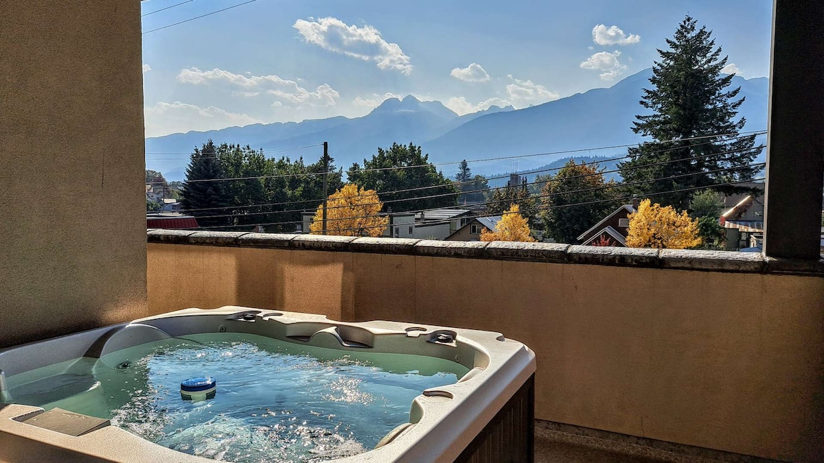 A hot tub sits on the balcony, offering views of distant mountains and trees in shades of green and gold. The clear sky is dotted with soft clouds, creating a relaxing atmosphere for outdoor enjoyment.
