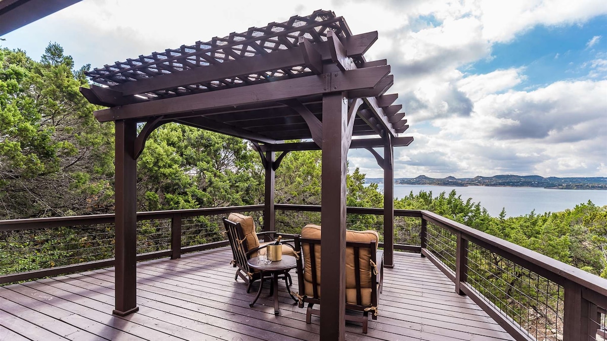 A shaded outdoor seating area features a pergola, providing a cozy space to relax. Two armchairs are positioned around a small table, with trees framing a scenic view of the lake in the background, complementing the tranquil atmosphere.