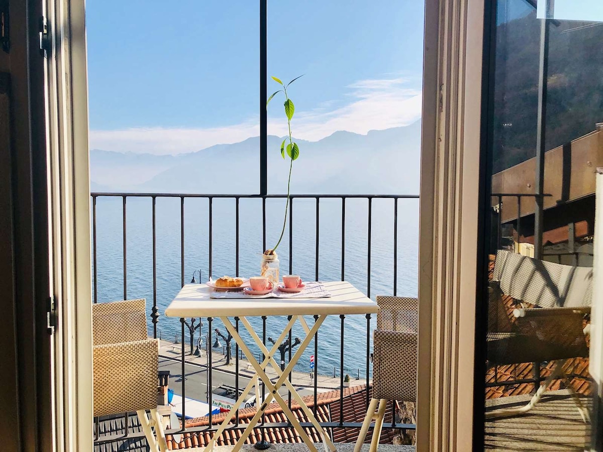 A small outdoor table is set for two, featuring pastries and cups. The view overlooks a serene lake, framed by distant mountains. The scene is enhanced by natural light streaming through the open door, creating an inviting atmosphere on the balcony.