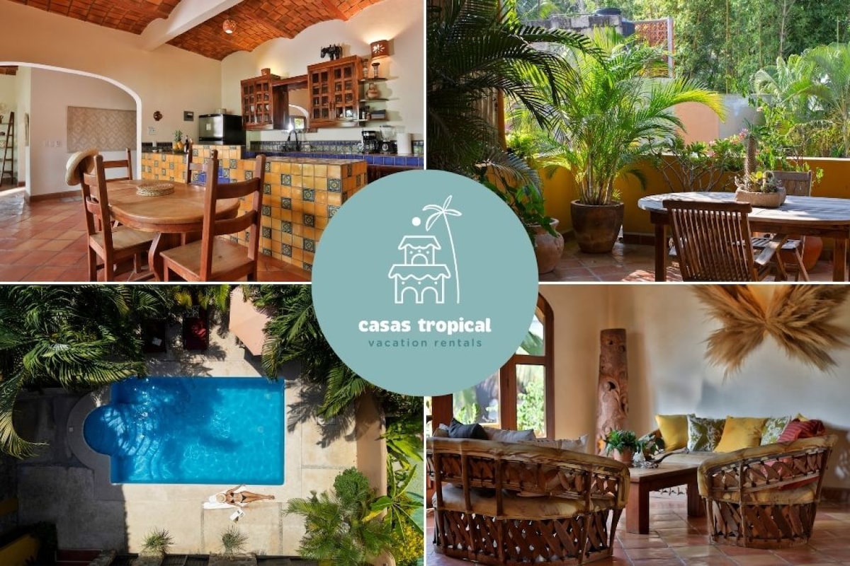 An inviting indoor space features a well-equipped kitchen with colorful tile accents and a dining area with a rustic wooden table and chairs. Lush greenery surrounds an outdoor patio area furnished with comfortable seating. A clear view of the tranquil pool invites relaxation.