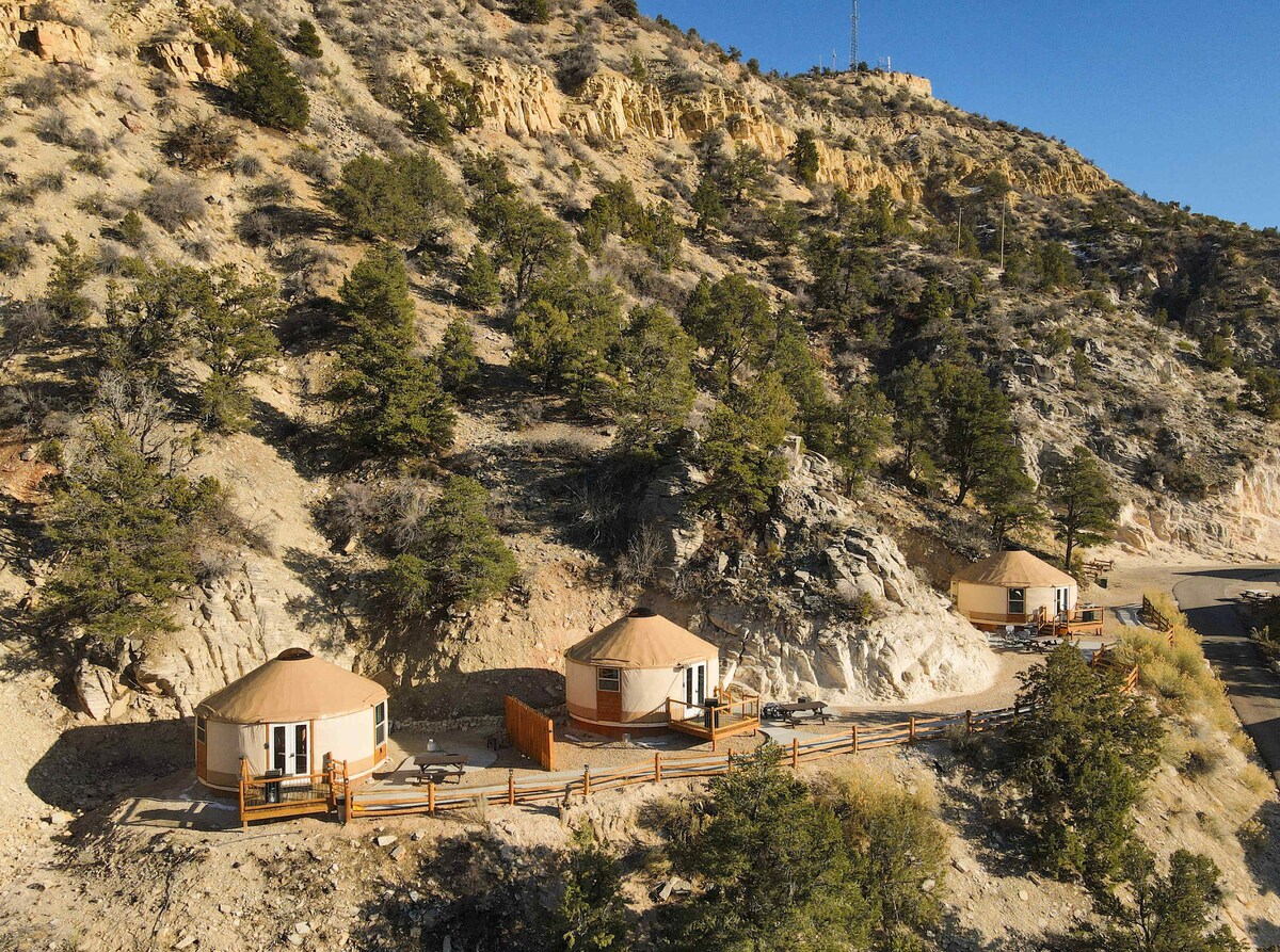 Three yurts are positioned along a hillside, surrounded by rugged terrain and trees. The natural landscape showcases a mix of rocky features and greenery, with a clear blue sky above. Each yurt has a wooden deck and large windows for views of the surroundings.