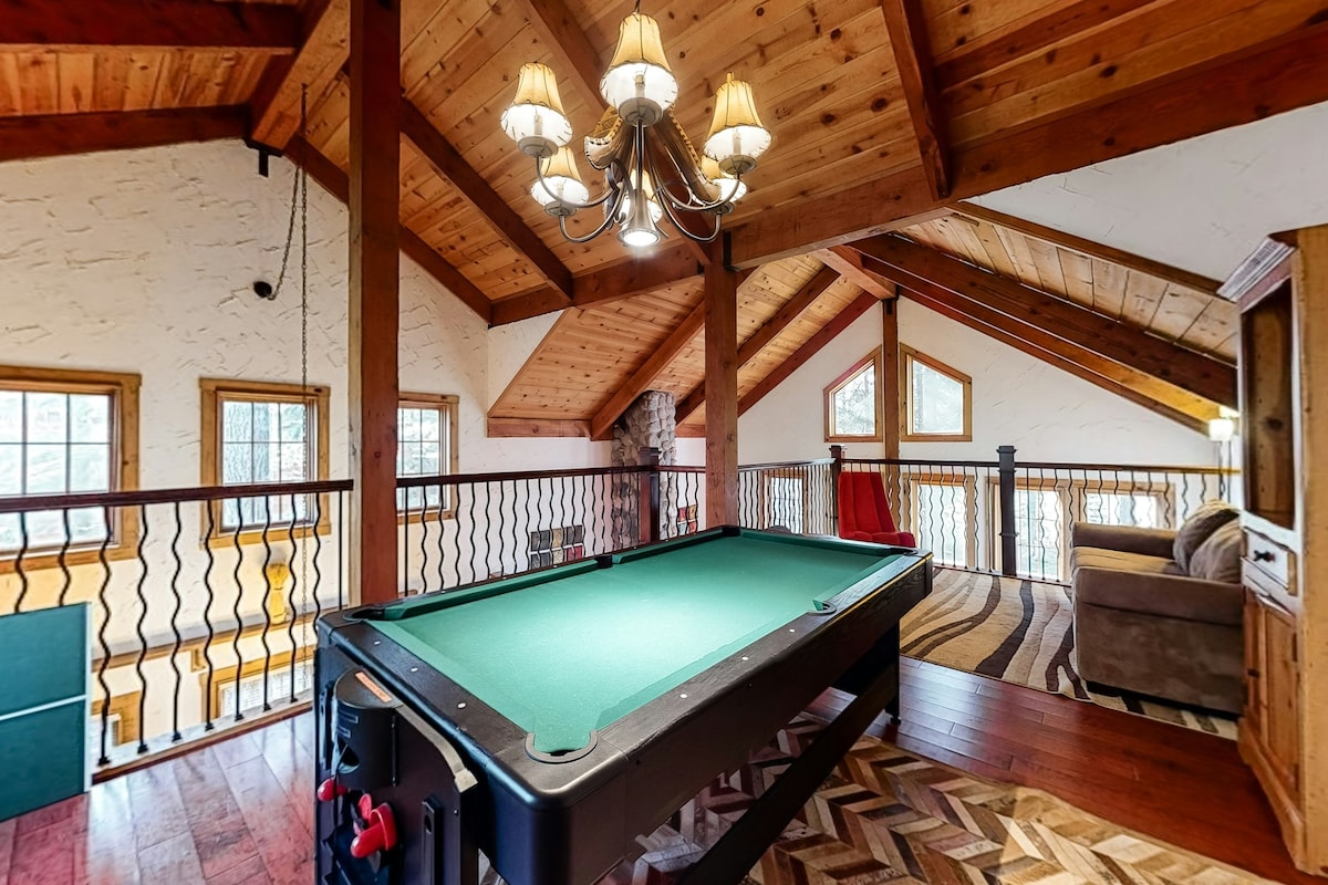 An inviting game area features a pool table beneath a chandelier with warm lighting. Natural wood beams accent the high ceiling, and windows allow filtered light to illuminate the room. A plush area rug adds texture, while a stone fireplace is visible in the background.