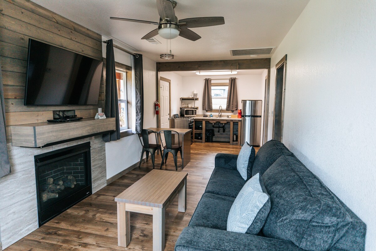 The living area features a comfortable dark gray sofa paired with a wooden coffee table. A mounted television hangs above a gas fireplace. Light pours in through a window adorned with gray curtains, while a dining table and kitchen area are visible in the background.