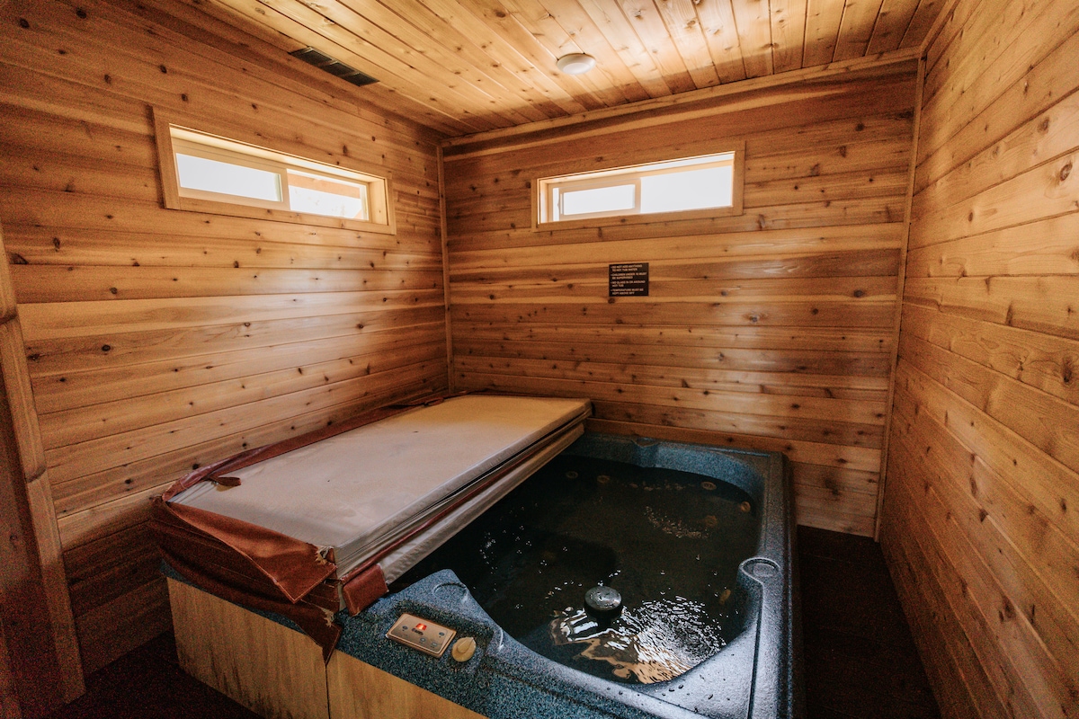 A private hot tub is housed in an exterior, attached room featuring wood-paneled walls. Natural light enters through small windows, creating a tranquil space for relaxation. The hot tub is equipped with a cover and controls, providing an inviting atmosphere for a soothing soak.