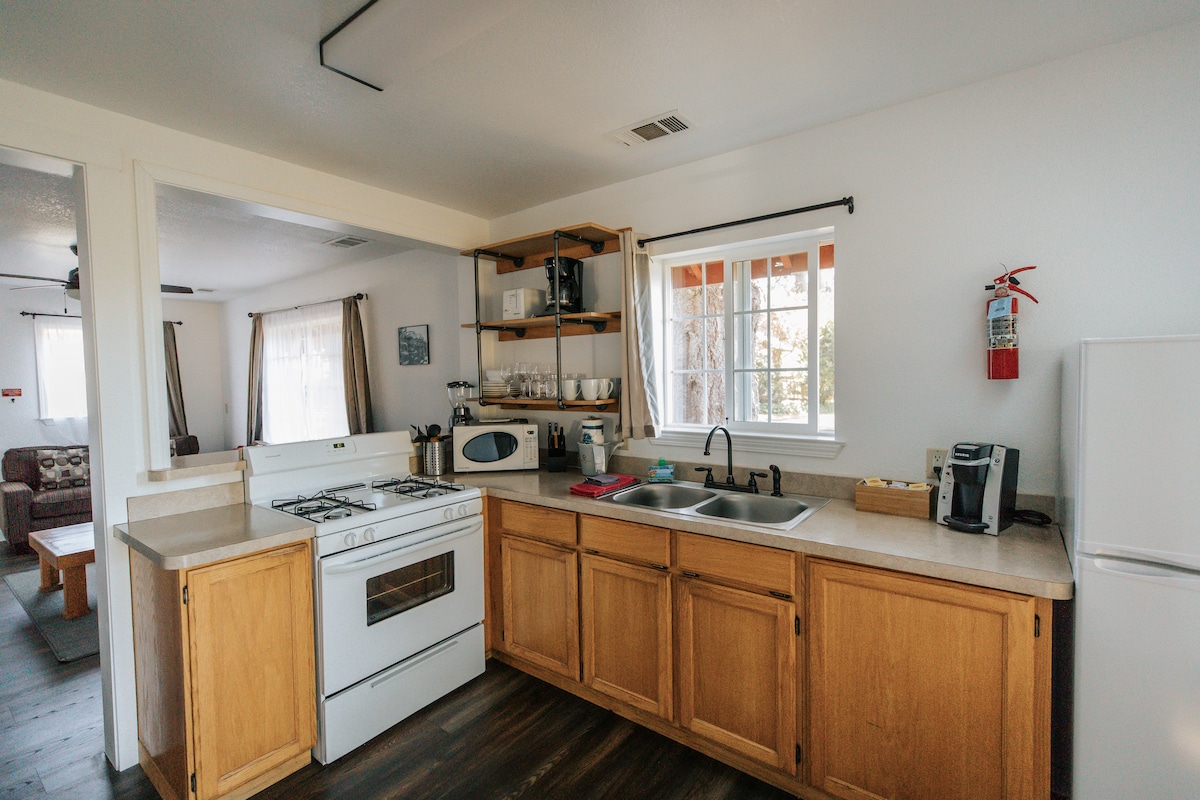 A fully equipped kitchen is presented, featuring ample cabinetry and a clean countertop. Appliances include a four-burner stove, oven, microwave, and refrigerator. A window above the sink allows natural light to filter in, while dishware is neatly arranged on shelving above.