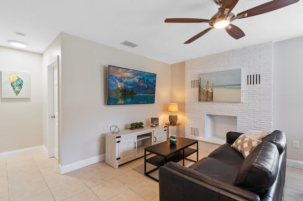 The living room features a sleek leather couch facing a mounted TV. A light-colored wooden entertainment unit provides storage, with a decorative lamp placed beside it. Wall art is displayed on adjacent walls, along with a white brick fireplace adding a touch of texture to the space.