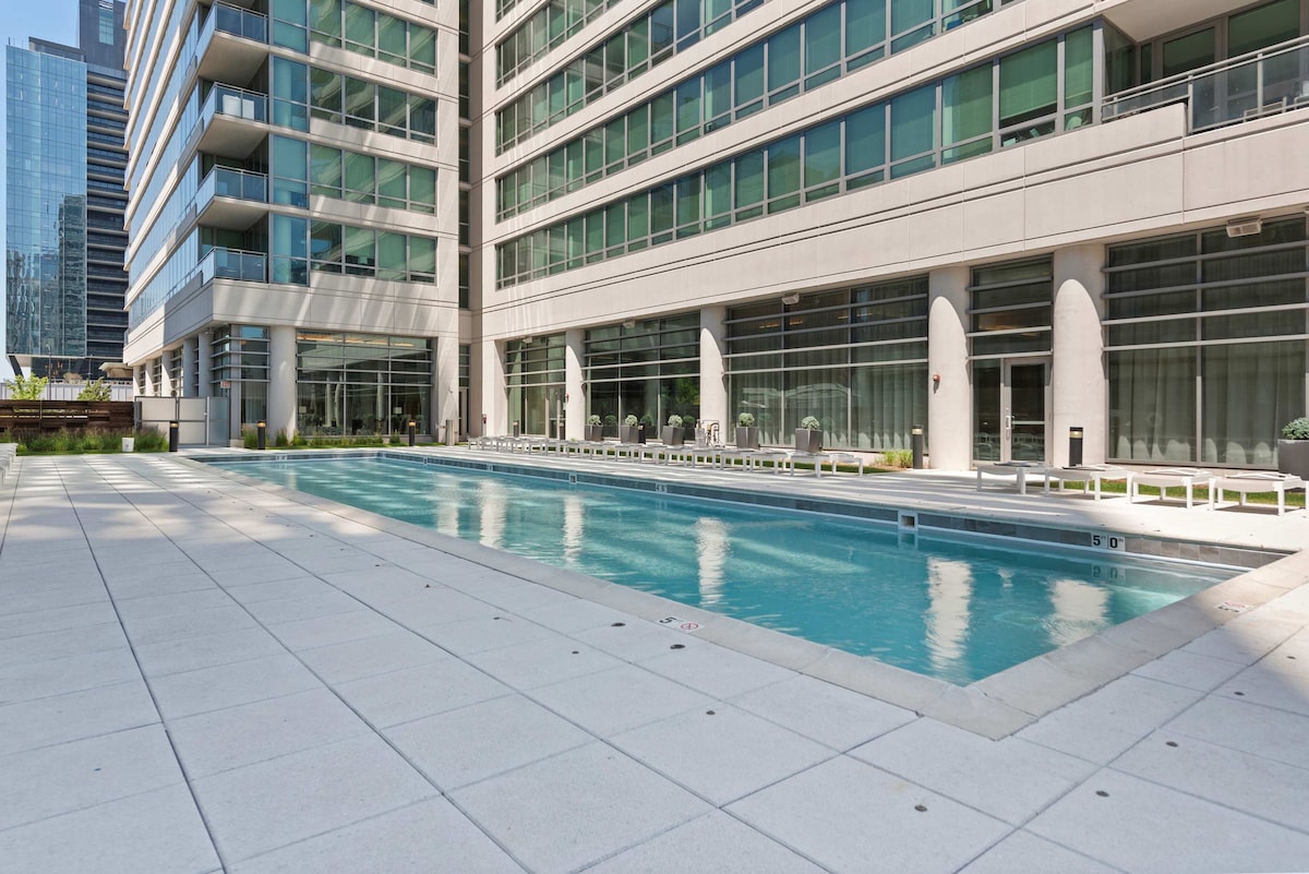An outdoor pool is viewed surrounded by modern building architecture. Lounge chairs are placed nearby on a spacious deck, and the pool's surface reflects sunlight. Tall glass windows of the building create a bright and open atmosphere.