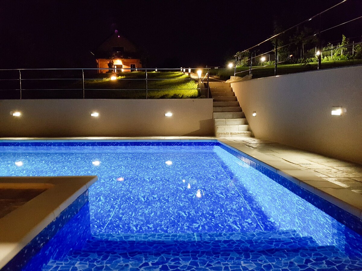 The illuminated swimming pool reflects a soft blue hue against the tiled surface, with steps leading up to a grassy area. Landscape lights guide the path, enhancing the peaceful evening atmosphere. The property is nestled in a tranquil countryside setting.