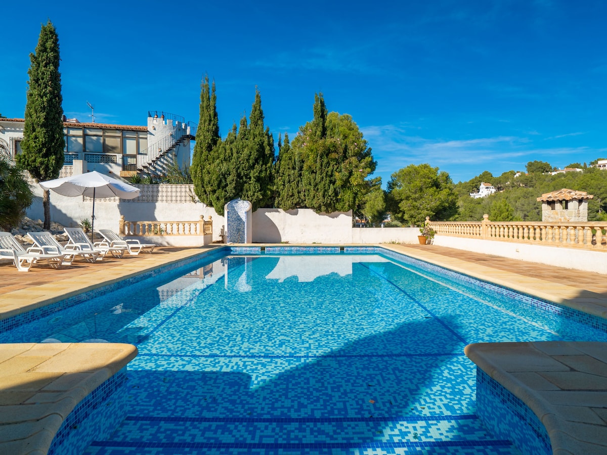 A large swimming pool is surrounded by a sun-drenched terrace, featuring loungers and umbrellas for relaxation. Lush greenery and a stone wall can be seen in the background, while a clear blue sky enhances the serene outdoor setting.