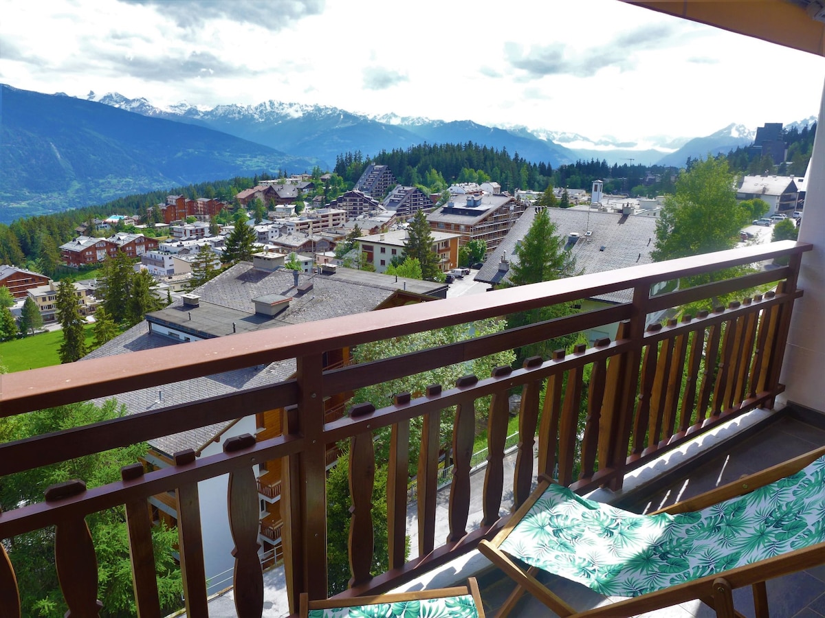 A spacious balcony features wooden railings and two deck chairs with green patterned fabric. The view includes a panorama of mountains and the surrounding countryside, showcasing a mix of residential buildings and greenery.