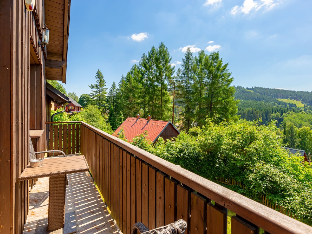 A wooden balcony offers views of lush greenery and distant hills. Two seating chairs provide a space to relax outdoors, while the surroundings feature a mix of trees and rustic rooftops, enhancing the tranquil atmosphere.