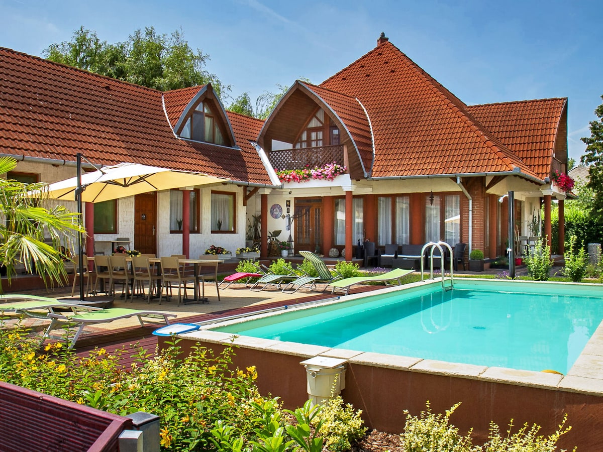 An inviting outdoor area is highlighted by a swimming pool and a patio with lounge chairs. A covered dining table accommodates several guests. The house features a distinct architectural design with a red tiled roof and blooming flower boxes, surrounded by well-maintained gardens.