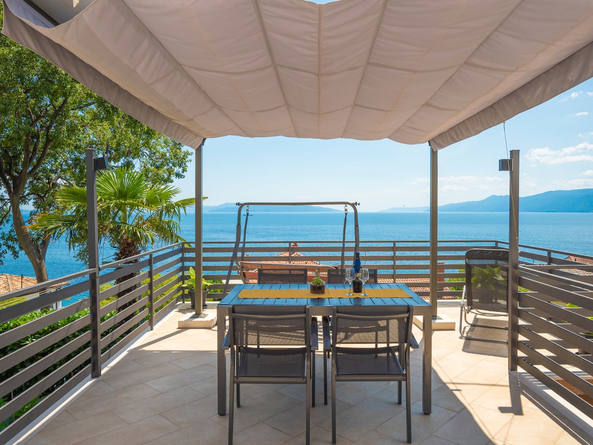 An outdoor terrace is presented with a table and chairs, overlooking a scenic view of the sea. A shaded area is provided by a fabric canopy, and a swing seat is situated in the background. Lush greenery borders the space, enhancing the serene environment.