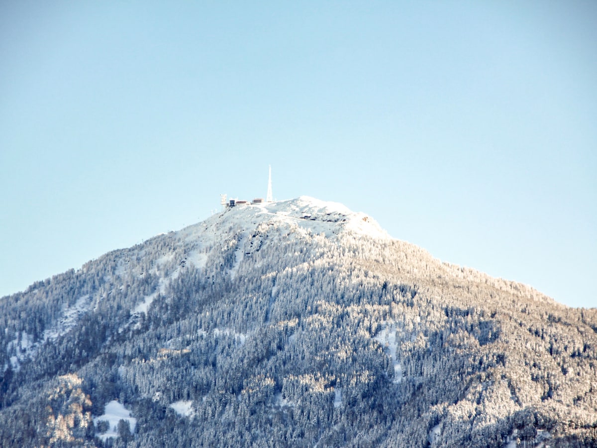 A snow-covered mountain peak is visible against a clear blue sky. The mountain's trees are blanketed in white snow, creating a serene contrast with the sky. A structure is perched atop the peak, adding interest to the natural landscape.