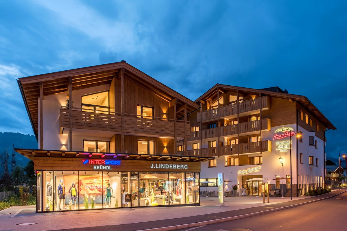 The modern alpine building features a wooden facade and multiple balconies, blending seamlessly with the surroundings. Illuminated shop fronts showcase sportswear, while soft evening light casts a welcoming glow over the entrance, highlighting the inviting atmosphere of the establishment.