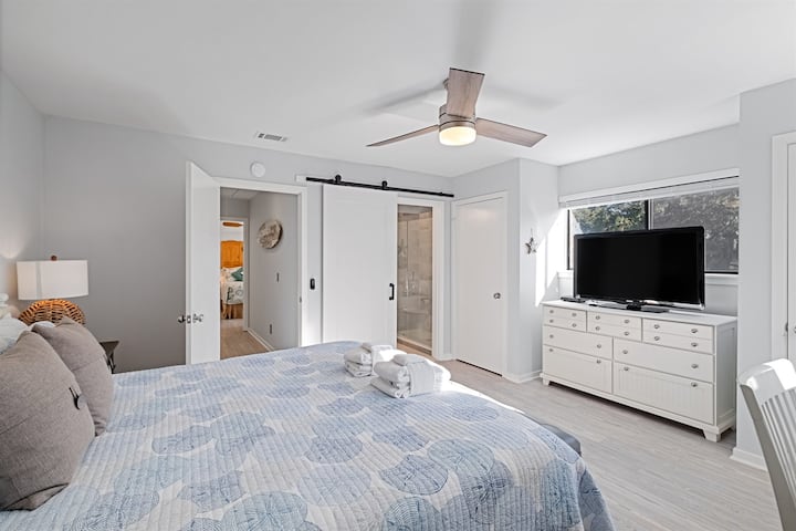 Chambre principale lumineuse et aérée à l'étage : lit matelassé, ventilateur de plafond, commode et porte coulissante de grange à la salle de bain attenante : des détails réfléchis pour une intimité ultime et une facilité d'accès à l'île.
