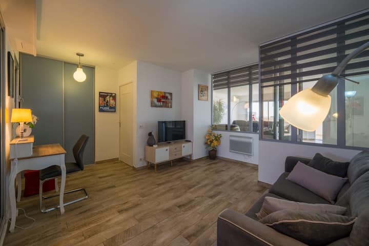 Salon lumineux avec coin bureau, parquet clair, canapé gris, TV sur meuble bas et grandes fenêtres à stores. Décoration moderne et sobre, éclairage chaleureux avec plusieurs lampes.