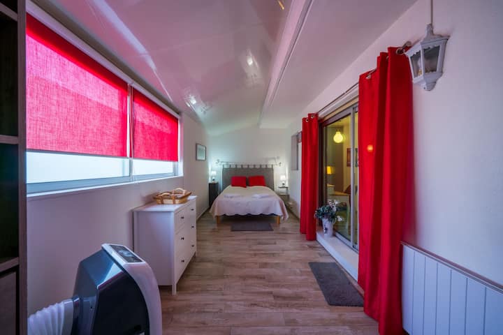 Chambre étroite et lumineuse avec lit double au fond, rideaux rouges marqués, commode blanche à gauche et baie vitrée à droite. Sol imitation bois, ambiance simple et cosy.