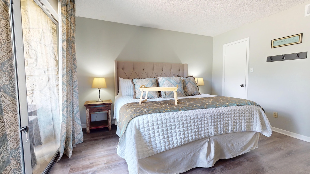 A well-appointed bedroom features a spacious upholstered bed adorned with decorative pillows. Soft lighting is provided by two bedside lamps on wooden tables. Natural light enters through large glass doors, which lead to an outdoor area, creating a calm and inviting atmosphere.