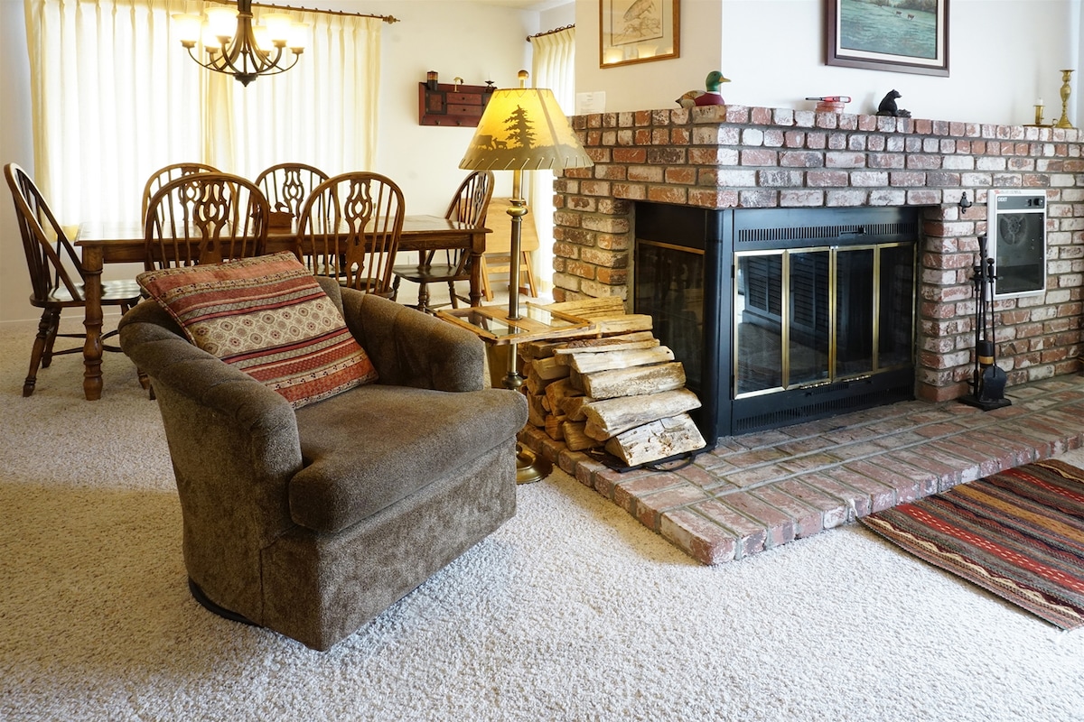 A comfortable seating area features a plush armchair next to a brick fireplace, which is stacked with firewood. A light-filled dining area is visible, showcasing a large wooden table surrounded by matching chairs, with soft drapes filtering sunlight.