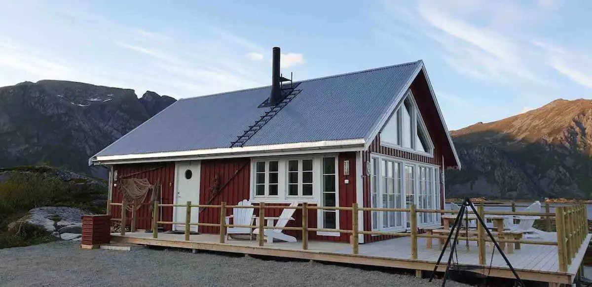 A red holiday cabin is depicted with a sloped metal roof and large panoramic windows. A spacious wooden deck surrounds the cabin, featuring white lounge chairs and a swing set. Breathtaking mountains rise in the background, complementing the serene outdoor space.