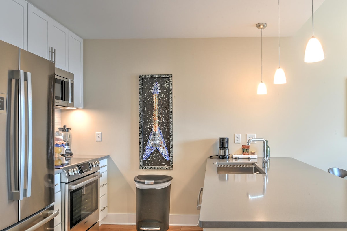 A modern kitchen is presented with stainless steel appliances, including a refrigerator and oven. Light fixtures hang above a spacious countertop, which is complemented by a mosaic art piece featuring a guitar. A trash can and coffee maker are visible, adding functionality.