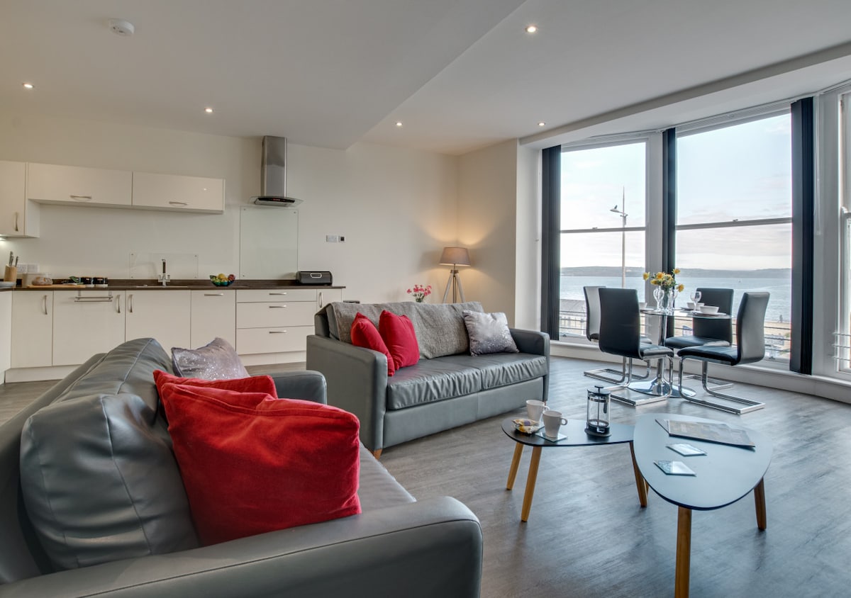 A modern living area features a bright, open layout with large windows showcasing sea views. Comfortable seating is arranged around a small coffee table, while a dining table for two sits near the kitchen. Natural light floods the space, accentuating the sleek design.