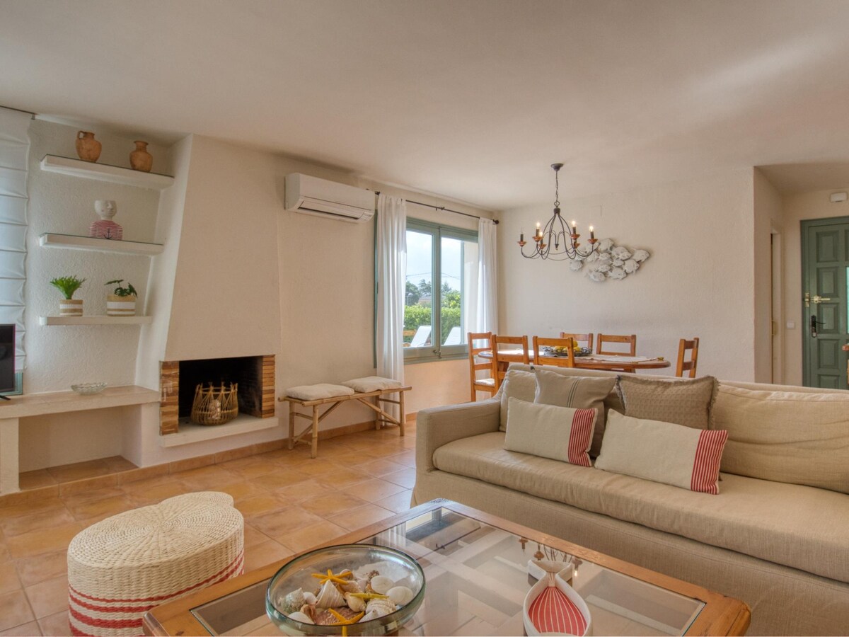 A bright living and dining area features a comfortable sofa adorned with various throw pillows. A dining table with wooden chairs sits nearby, illuminated by natural light from the window. A fireplace is positioned against a textured wall, providing warmth and a relaxing focal point.