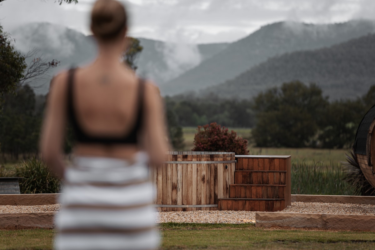 A scenic outdoor area featuring a wooden hot tub, surrounded by gravel, with a backdrop of rolling hills and misty mountains. Lush greenery is visible in the foreground, enhancing the serene atmosphere.