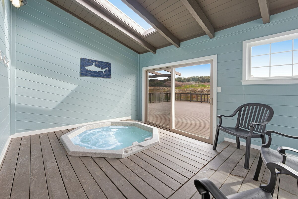An enclosed hot tub is prominently featured in a sunlit corner, surrounded by light blue walls. The area includes two black chairs, and large glass doors open to a spacious outdoor deck, providing a view of the coastline.