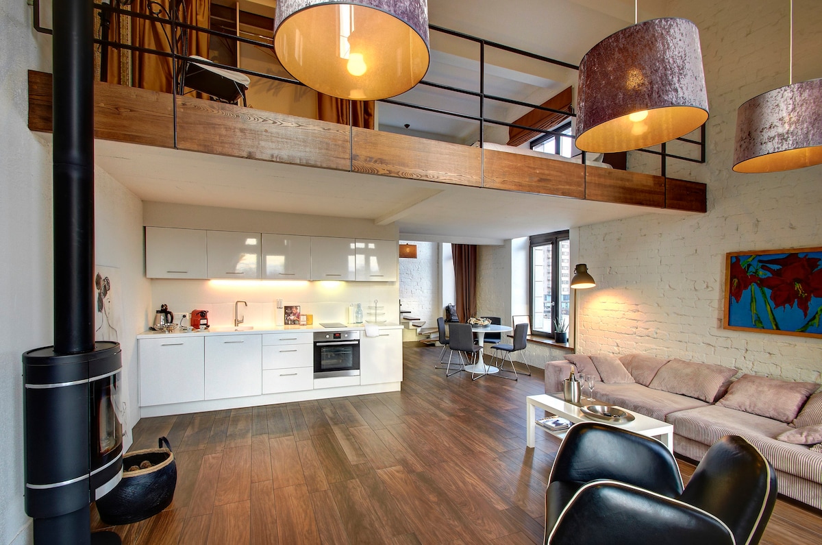 A spacious living area is showcased with a modern kitchen featuring a microwave and stovetop. A comfortable couch is positioned near a sleek black chair. Large windows allow natural light to brighten the space, while wooden beams add architectural interest above.