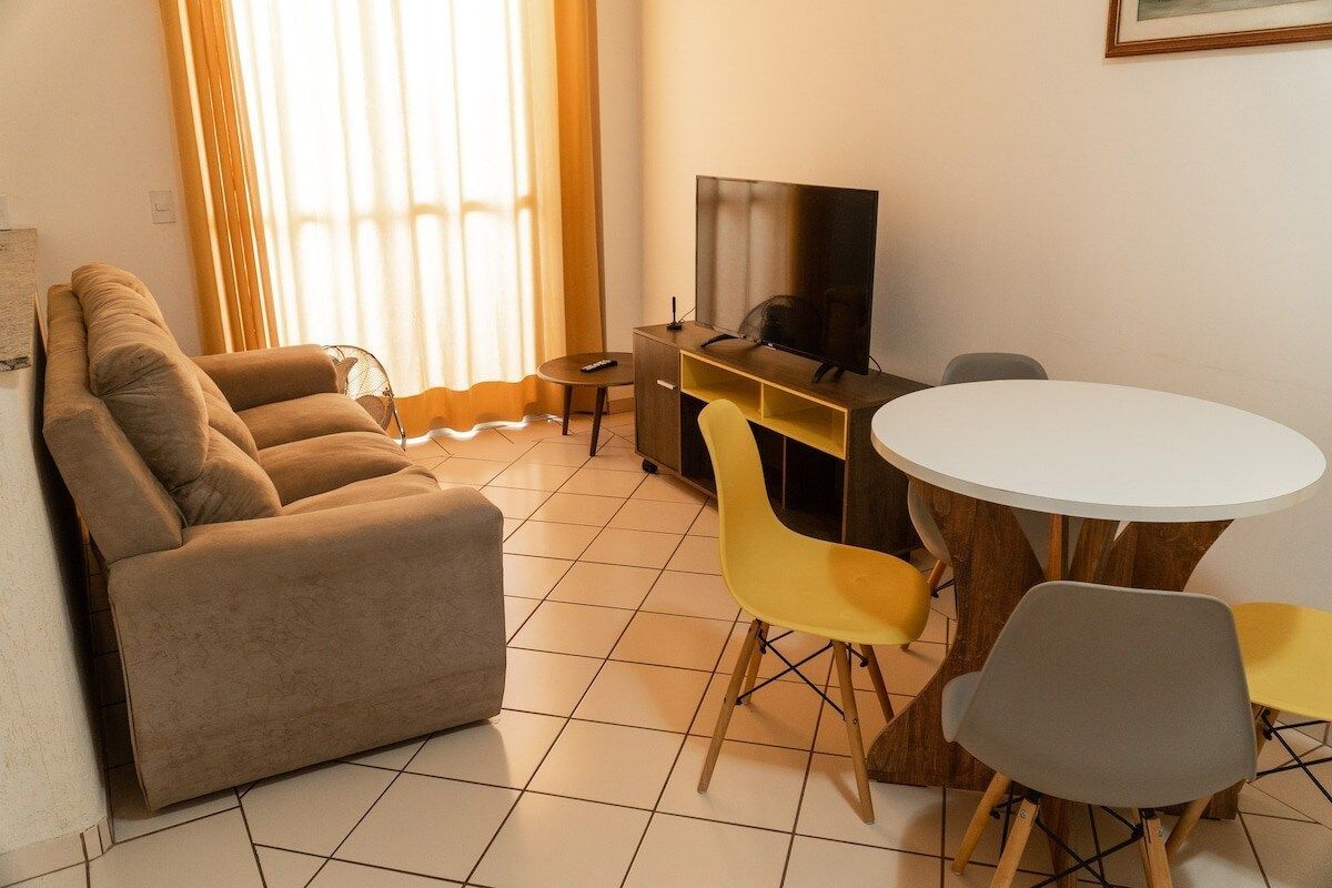 A living area features a spacious beige sofa and a round table surrounded by colorful chairs. A flat-screen TV sits on a stand, with natural light flooding in through floor-to-ceiling curtains, enhancing the bright and airy feel of the space.