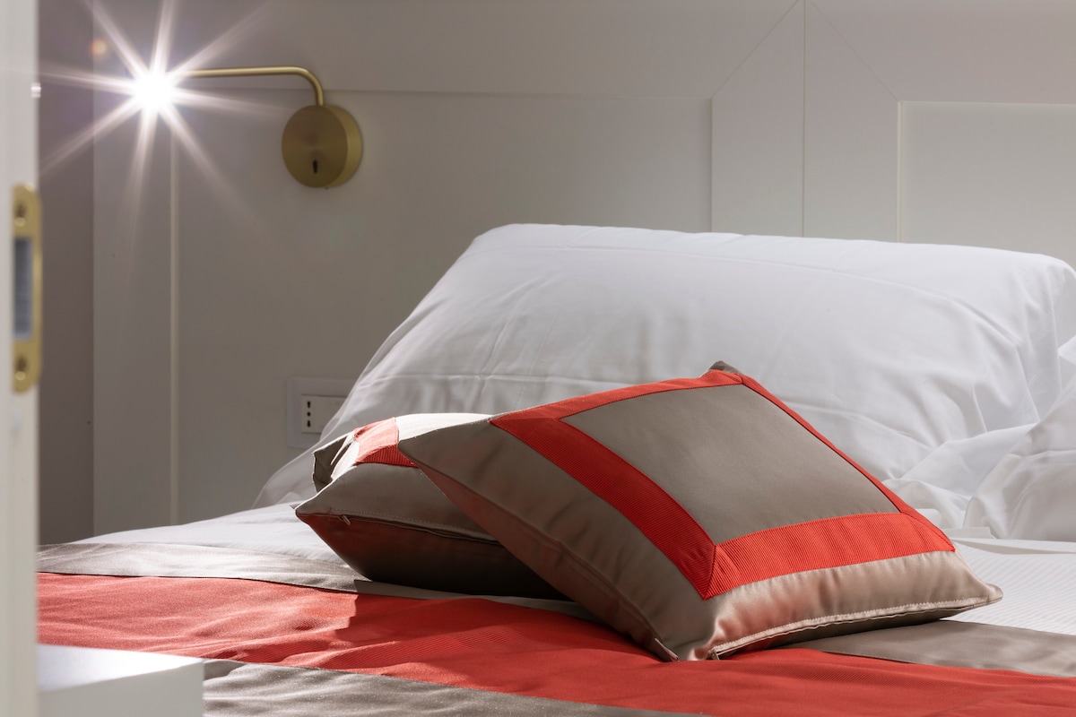 A spacious bed is dressed in soft white linens, featuring two decorative pillows with a geometric pattern in red and beige. A modern bedside lamp provides targeted lighting, enhancing the room's inviting ambiance.