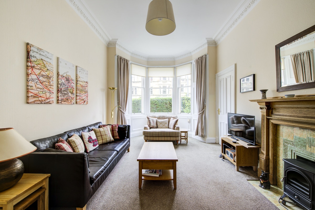 A spacious lounge area is featured with a dark leather sofa, a light-colored armchair, and a wooden coffee table. Natural light floods in through large bay windows, with beige curtains elegantly framing the view. A TV and decorative fireplace complete the inviting setting.