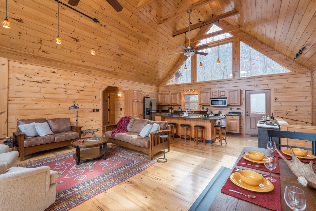 An open living space features high wooden ceilings and large windows allowing natural light to fill the room. A cozy seating area is arranged with a sofa and armchairs, accompanied by a dining space set for six. The fully equipped kitchen is visible in the background.