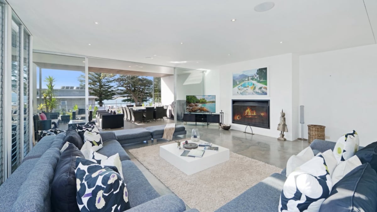 An open-plan living area features a large sectional sofa adorned with decorative cushions, arranged around a central coffee table. A soft rug anchors the seating, and large glass doors allow natural light to fill the space, with views of the outdoor dining area and pool.