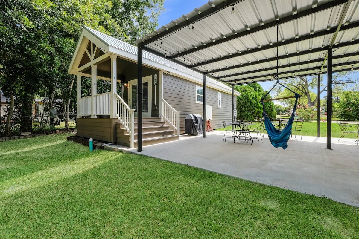 A spacious outdoor area features a covered patio with a hanging hammock, surrounded by lush green grass. The tiny home is adjacent, showcasing steps leading to the entrance. Several tables are set up for dining or relaxation under the expansive roof.