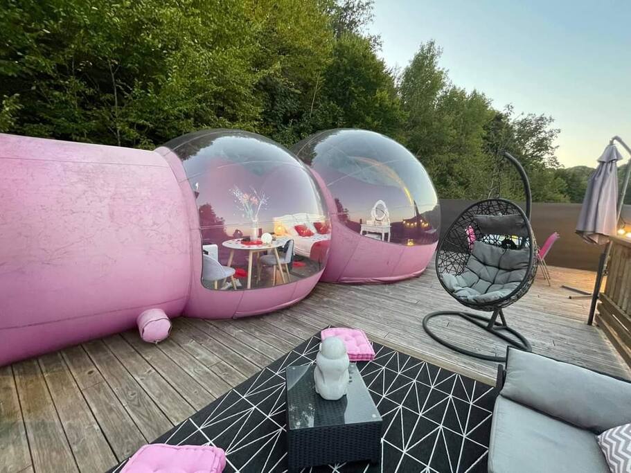 Two transparent spheres, colored in soft pink, are situated on a wooden deck surrounded by greenery. A modern hanging chair and outdoor seating area are visible, along with a table set for dining inside one bubble. The scene captures a serene outdoor setting at dusk.