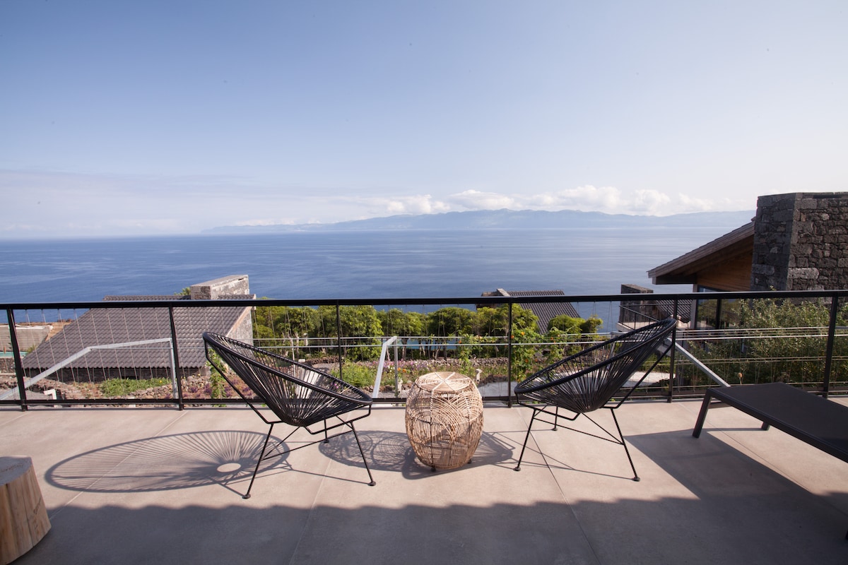 A large balcony features two black wicker chairs positioned to take in expansive ocean views. A natural wooden table stands nearby, while the horizon showcases the outline of São Jorge Island under a clear blue sky.