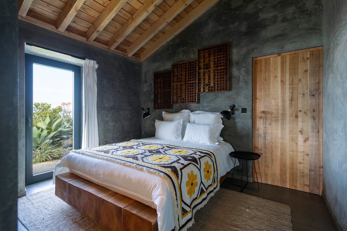 A spacious bedroom features a wooden bed frame topped with white pillows and a colorful patterned blanket. Concrete walls are complemented by wooden beams on the ceiling. A doorway opens to a view of greenery, and a small side table stands beside the bed.