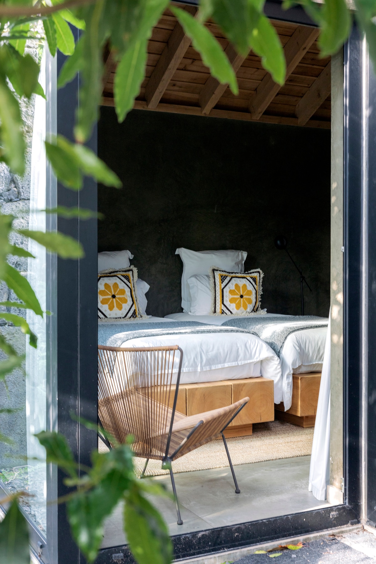 A cozy bedroom is framed by lush greenery, featuring two spacious beds adorned with decorative pillows. Natural light streams in through large glass openings, illuminating the wooden ceiling and soft bedding. A metal chair adds a touch of design to the inviting space.