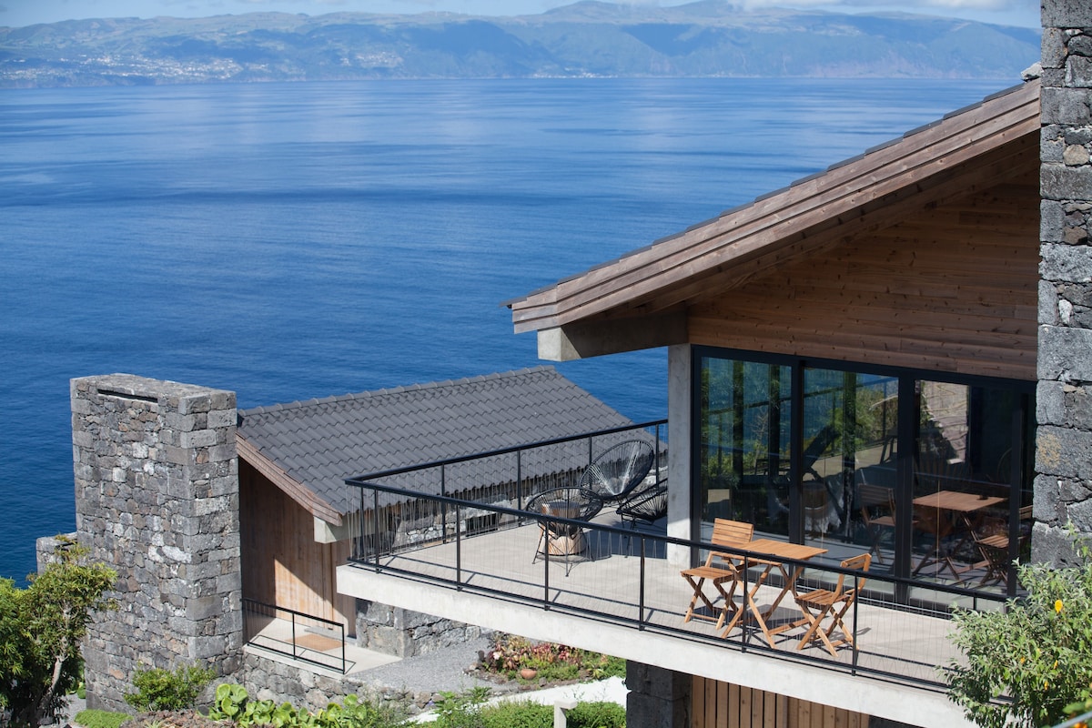 A spacious balcony extends from the house, featuring wooden furniture set against a backdrop of expansive ocean views. The structure combines stone and wood elements, with large glass windows reflecting the serene blue of the sea. Lush greenery frames the scene.