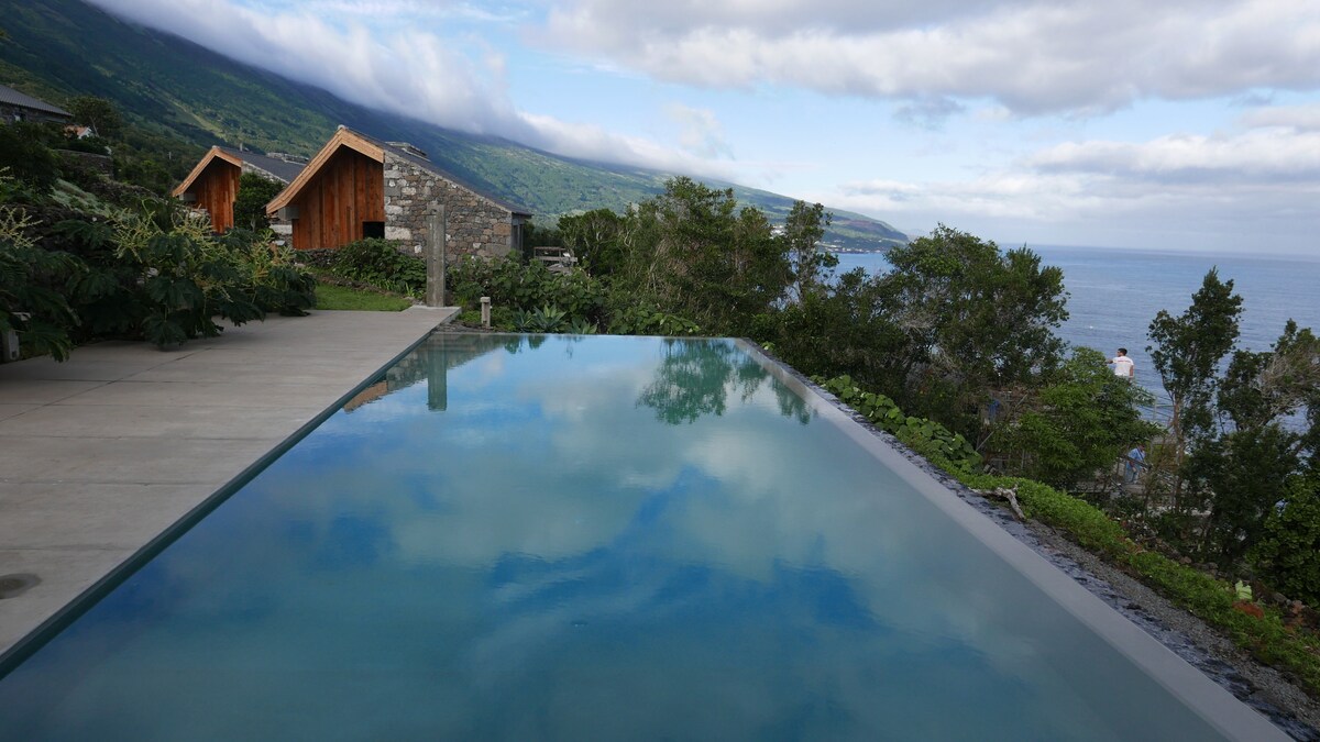 An infinity pool stretches across the foreground, reflecting the sky and surrounding greenery. In the background, two rustic cabins are nestled among lush vegetation, with a scenic ocean view and rolling hills under a partly cloudy sky.