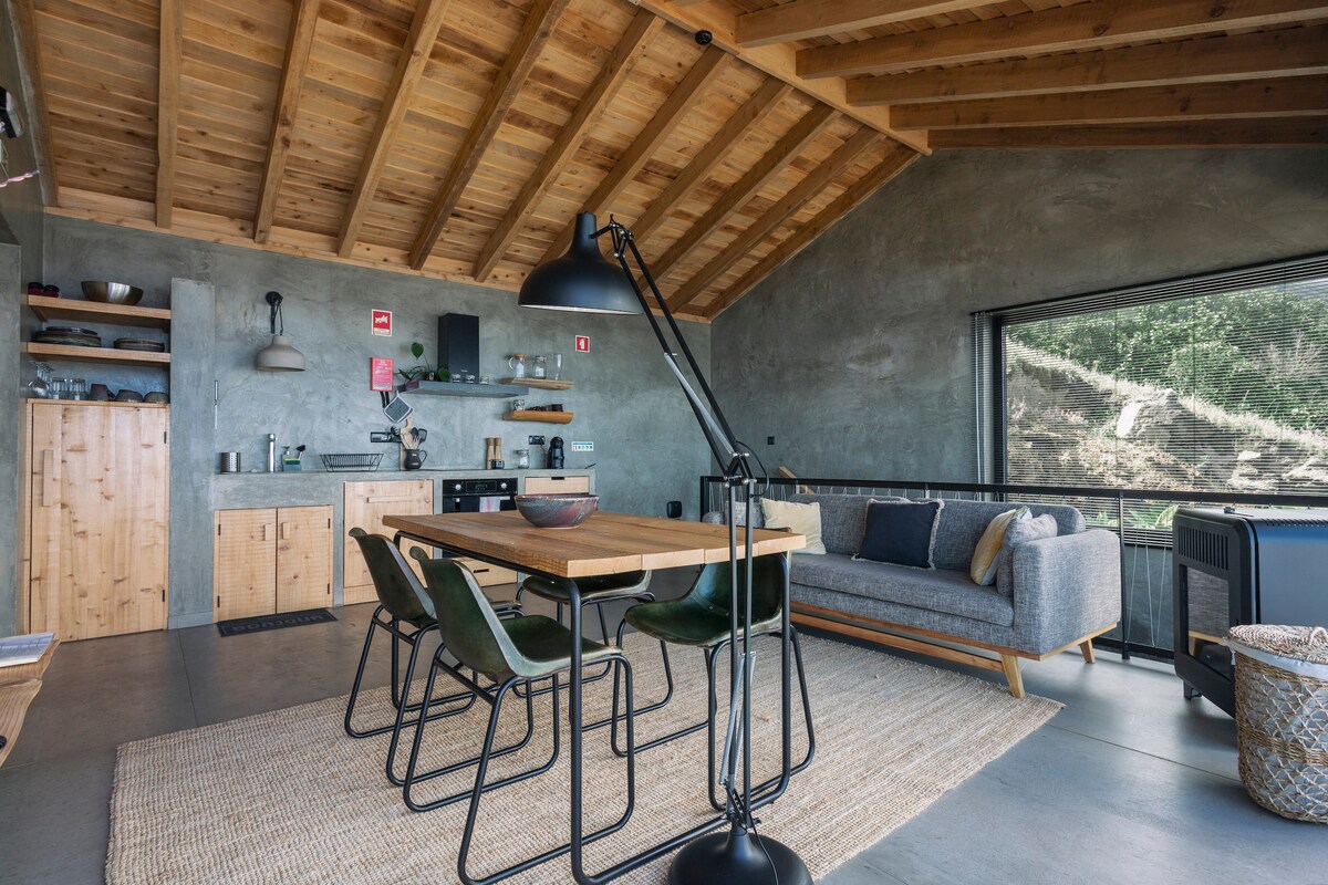 An open-plan living and dining area features a wooden ceiling and modern concrete walls. A large dining table is surrounded by chairs, with a sofa positioned near expansive glass sliding doors, offering views of the surrounding landscape. A fully equipped kitchen is visible in the background.