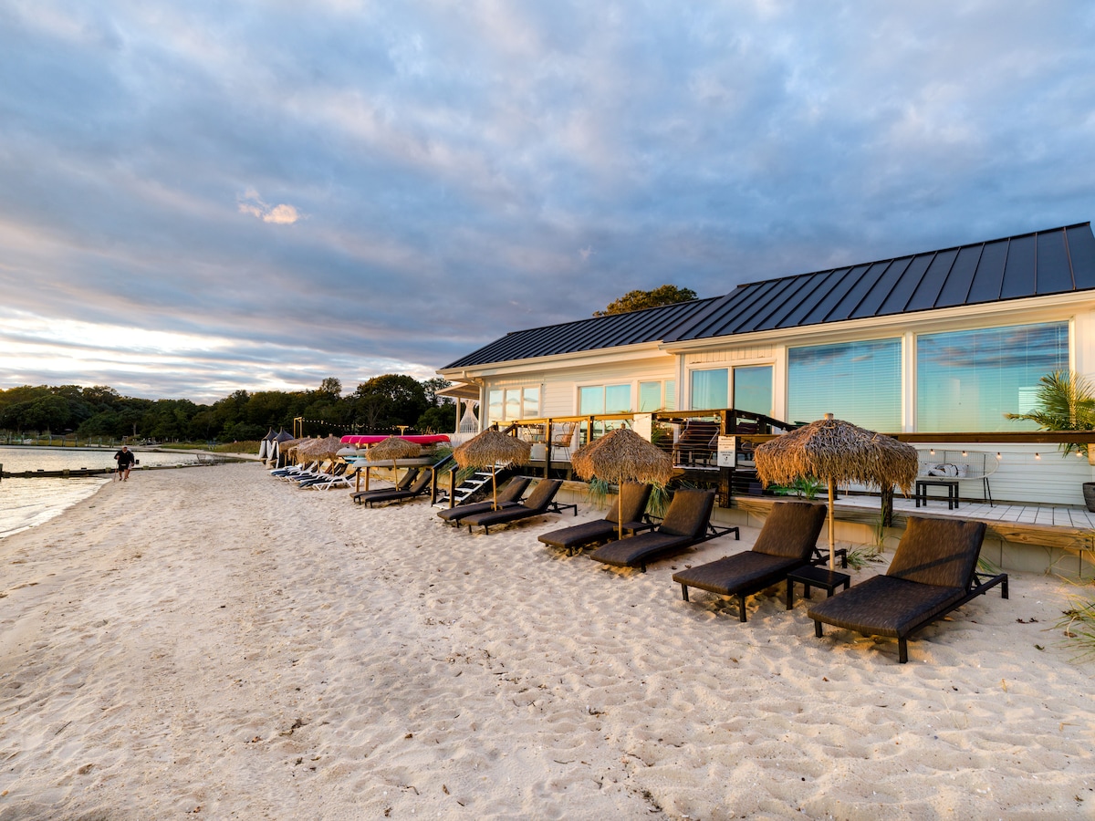 A sandy beach area is lined with several lounge chairs and umbrellas, facing the water. In the background, a building with large windows is visible, reflecting the sky and surrounding nature.